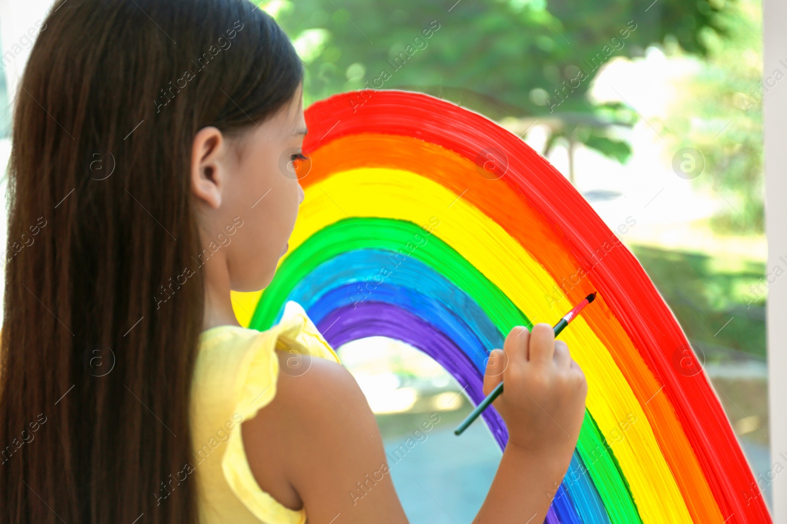 Photo of Little girl drawing rainbow on window indoors. Stay at home concept