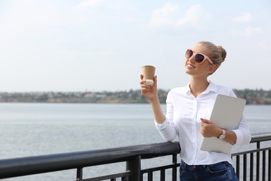 Young woman with cup of coffee and laptop at pier. Joy in moment