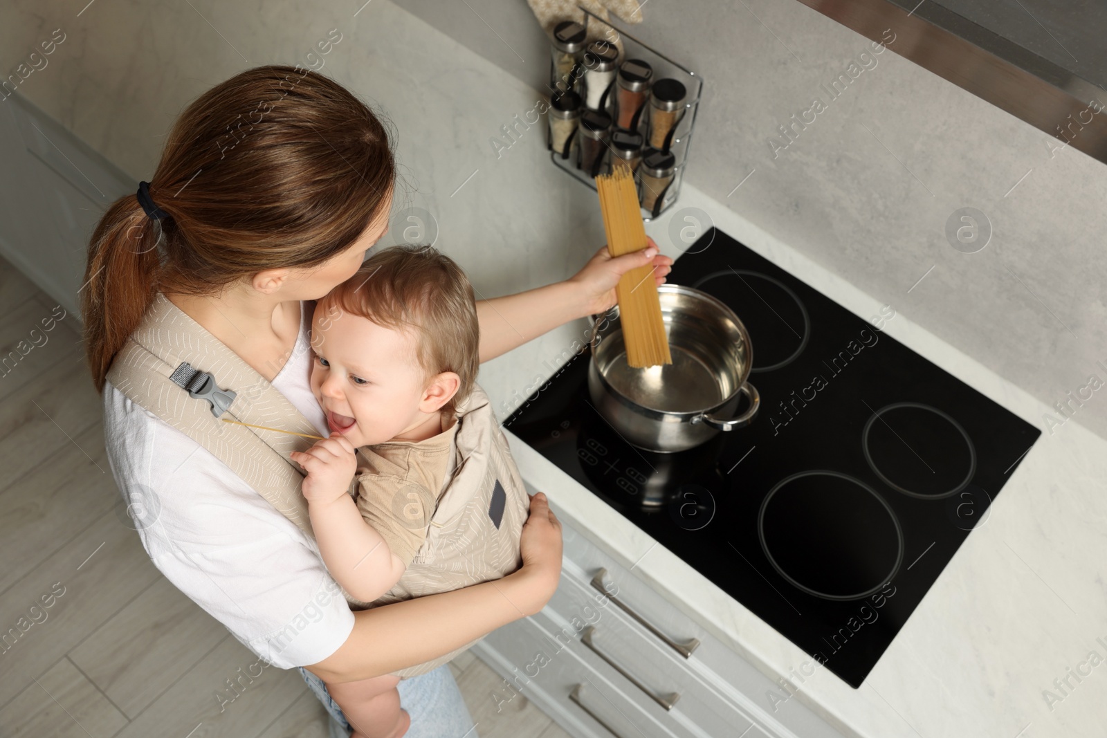 Photo of Mother holding her child in sling (baby carrier) while cooking pasta in pot indoors, above view