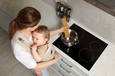 Mother holding her child in sling (baby carrier) while cooking pasta in pot indoors, above view