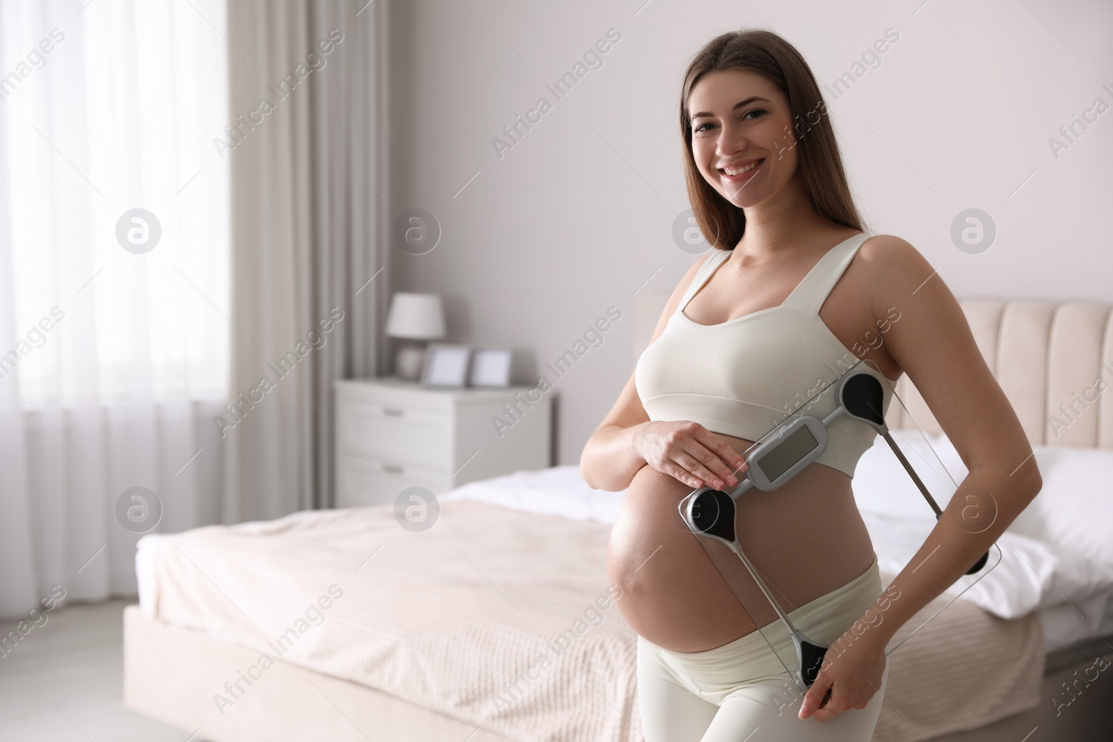 Photo of Young pregnant woman with scales in bedroom. Space for text