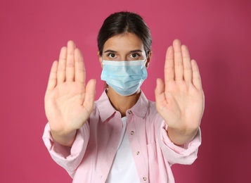 Photo of Woman in protective mask showing stop gesture on pink background. Prevent spreading of coronavirus