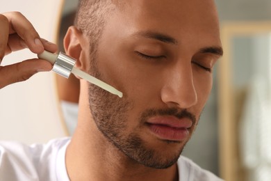 Photo of Handsome man applying cosmetic serum onto face indoors, closeup