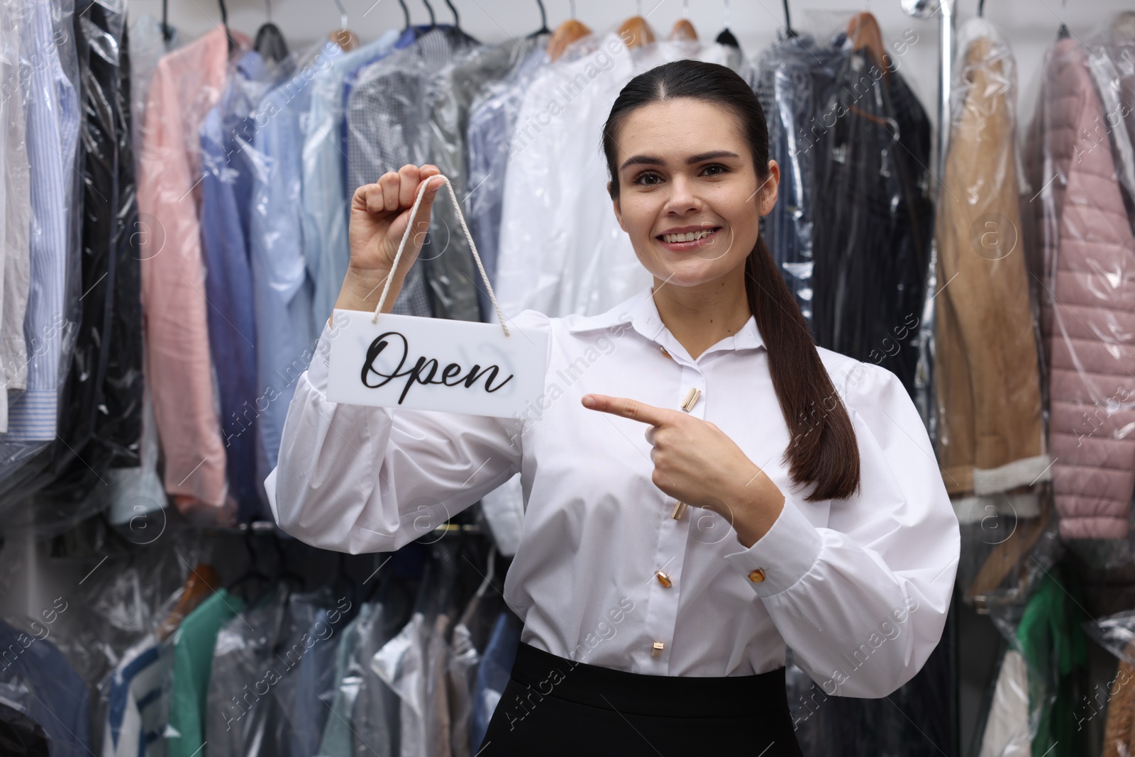 Photo of Dry-cleaning service. Happy worker holding Open sign near rack with clothes indoors