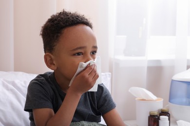 African-American boy with tissue blowing nose in bed indoors, space for text. Cold symptoms