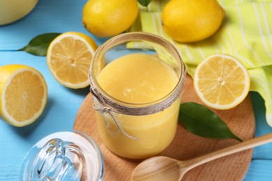 Photo of Delicious lemon curd in glass jar, fresh citrus fruits, green leaves and spoon on light blue wooden table