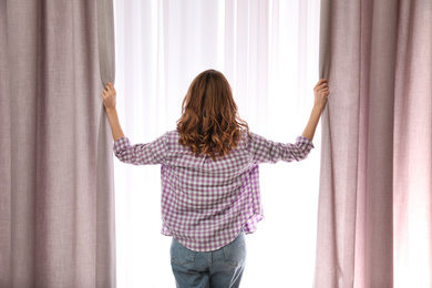 Woman opening window curtains at home in morning, back view