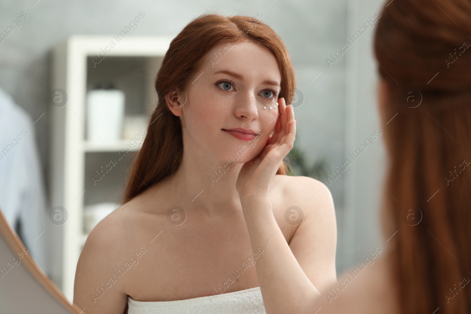 Photo of Beautiful woman with freckles and cream on her face near mirror in bathroom