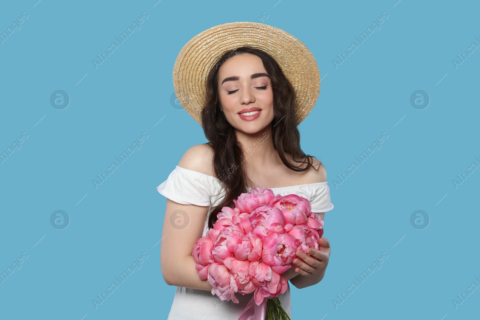 Photo of Beautiful young woman in straw hat with bouquet of pink peonies against light blue background