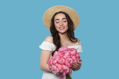 Photo of Beautiful young woman in straw hat with bouquet of pink peonies against light blue background