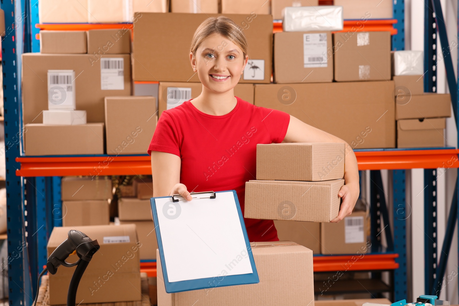 Photo of Post office worker with parcels and clipboard near rack indoors