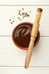 Photo of Tasty barbeque sauce in bowl, brush and peppercorns on white wooden table, top view