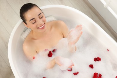 Photo of Happy woman taking bath in tub with foam and rose petals indoors, top view