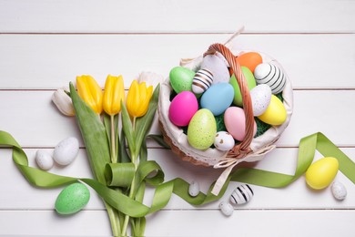 Easter eggs in basket, tulip flowers and ribbon on white wooden table, flat lay