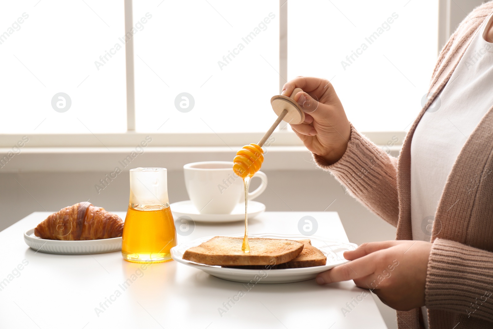 Photo of Woman pouring honey onto toast at white table, closeup