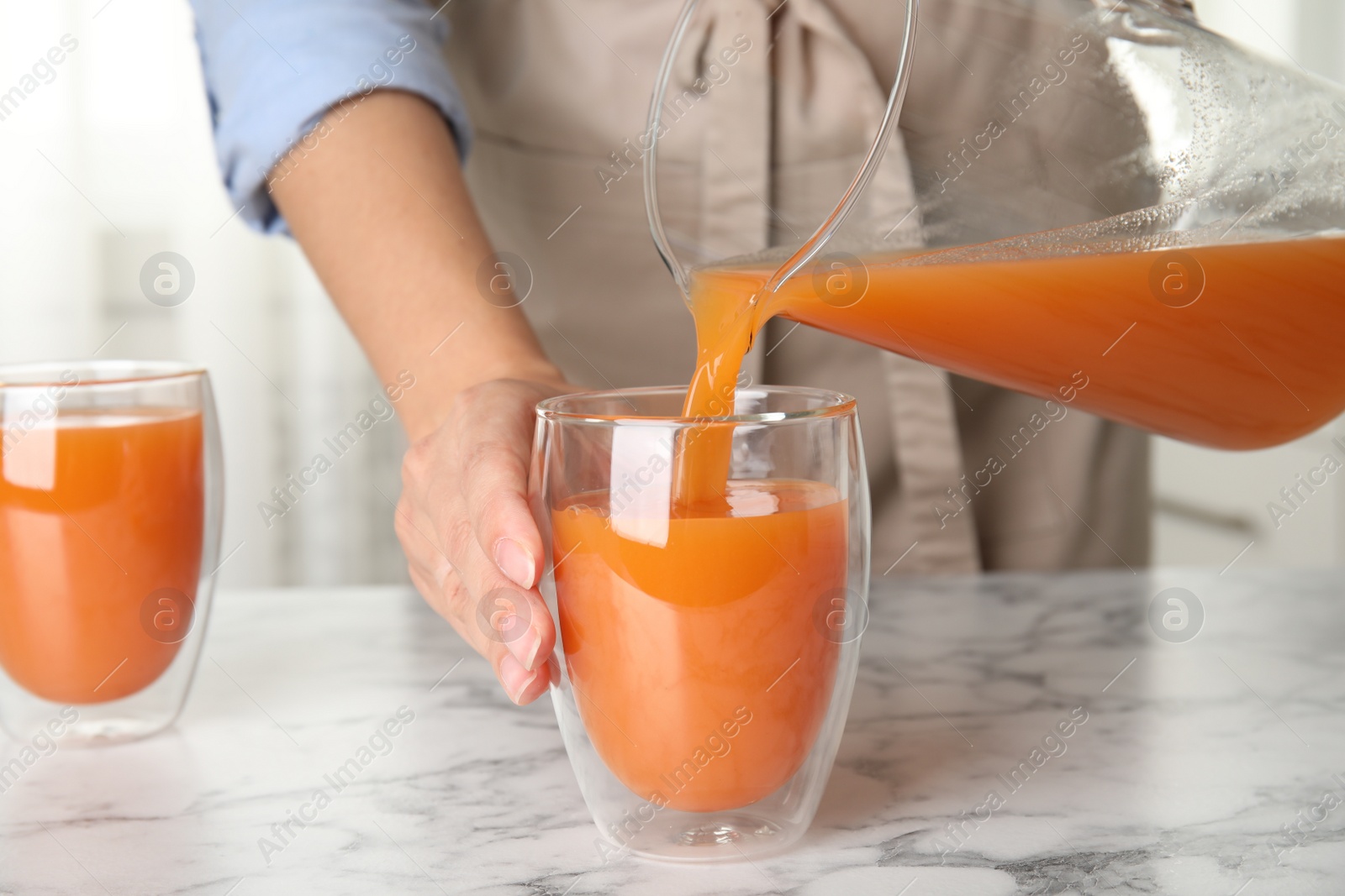 Photo of Woman pouring freshly made carrot juice into glass on white marble table in kitchen, closeup