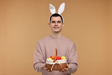 Photo of Easter celebration. Handsome young man with bunny ears holding basket of painted eggs on beige background