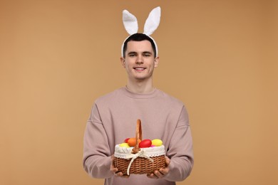 Easter celebration. Handsome young man with bunny ears holding basket of painted eggs on beige background