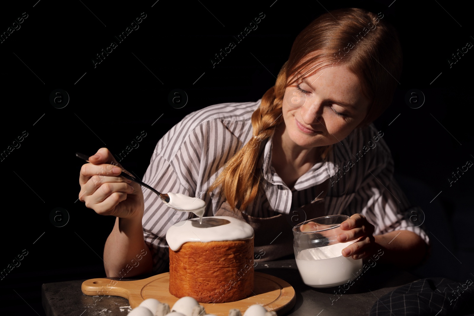 Photo of Young woman decorating traditional Easter cake with glaze at table against black background