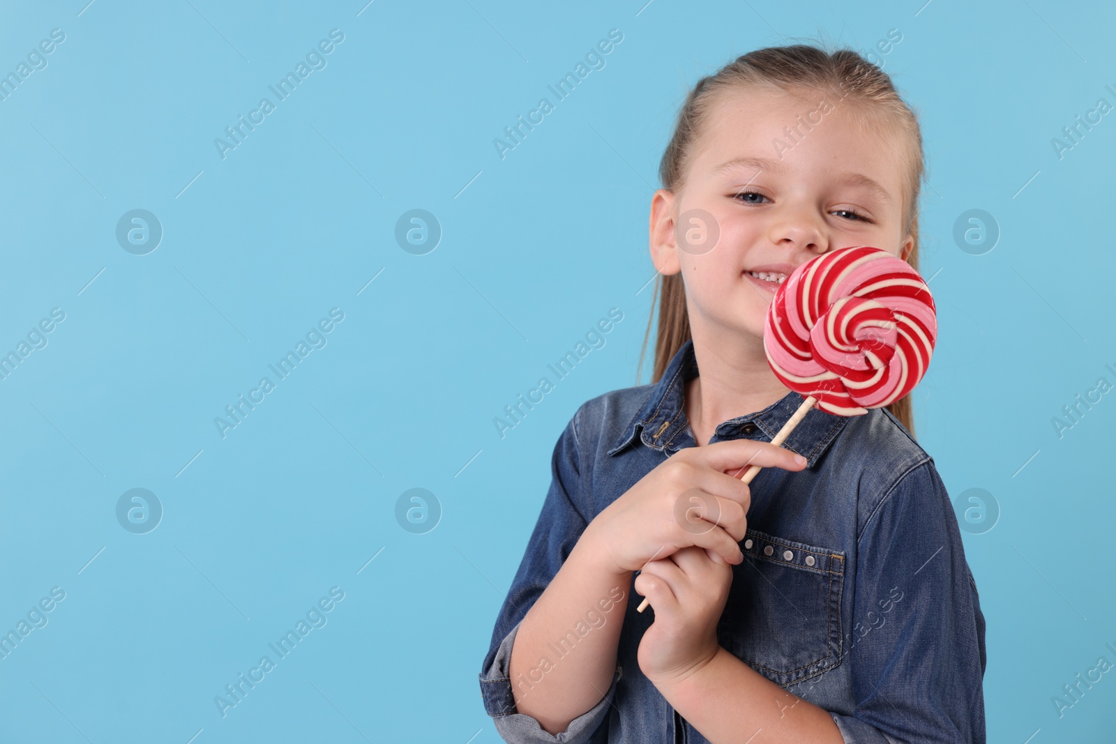 Photo of Happy little girl with bright lollipop swirl on light blue background, space for text