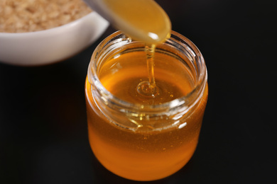 Photo of Spoon with honey over jar on black table, closeup