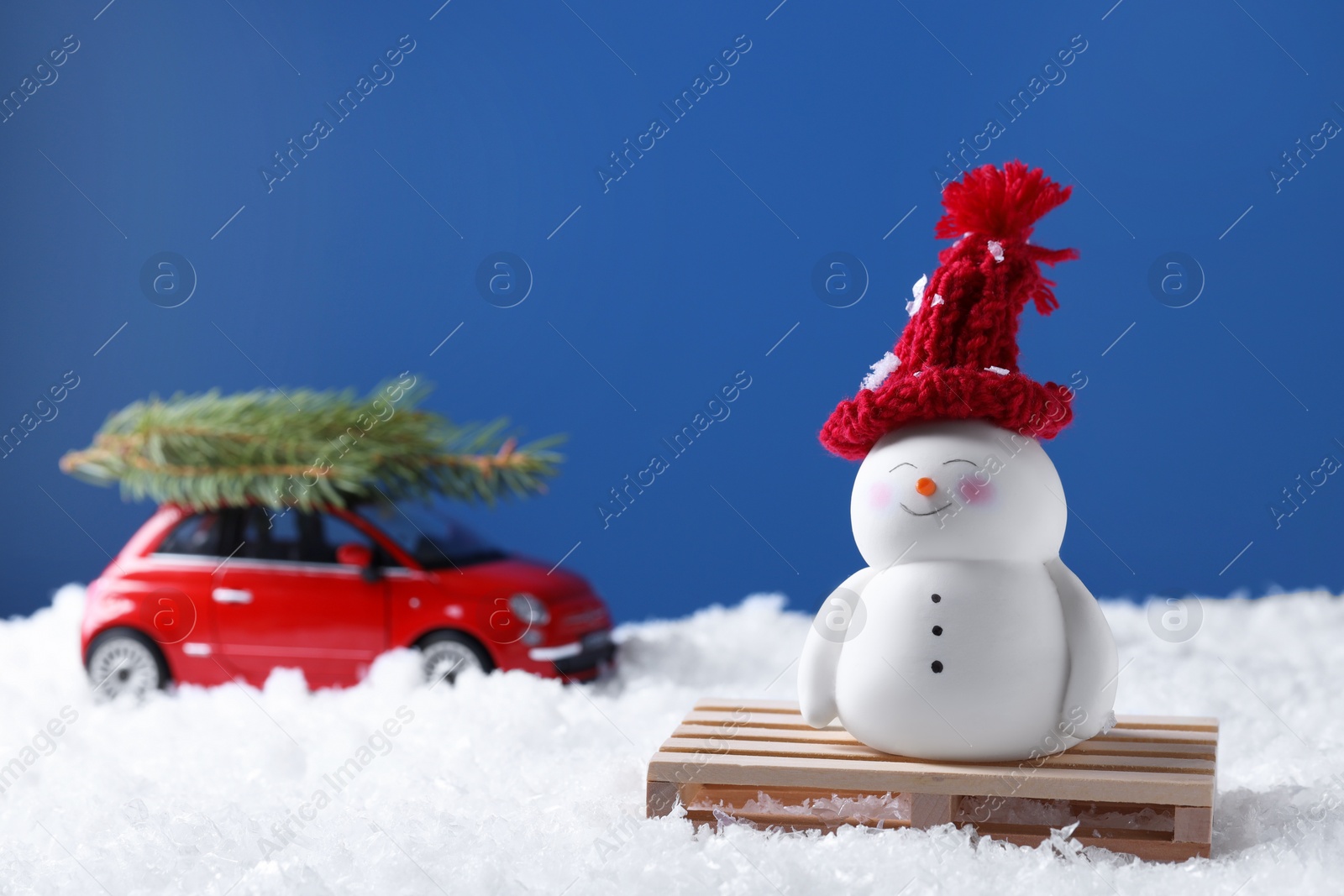 Photo of Cute decorative snowman and toy car with fir tree branches on artificial snow against blue background