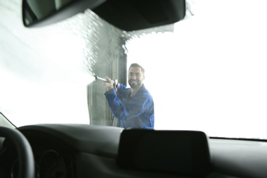 Worker cleaning automobile windshield with high pressure water jet at car wash, view from inside