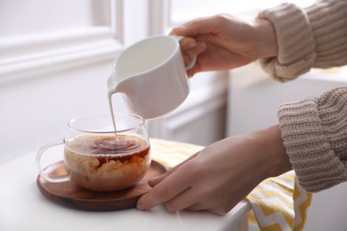 Woman pouring milk into cup of tea at white table, closeup