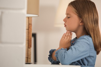 Photo of Cute little girl with hands clasped together praying at home. Space for text