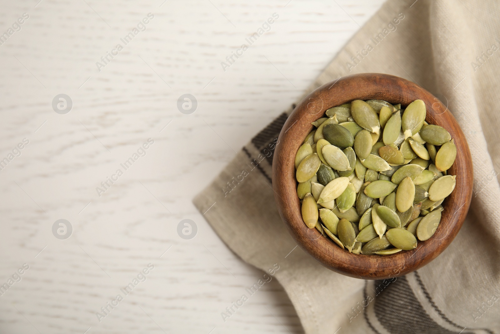 Photo of Bowl of pumpkin seeds with napkin on white wooden table, top view. Space for text