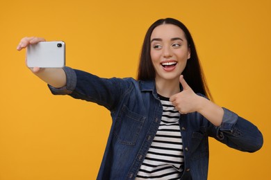 Photo of Smiling young woman taking selfie with smartphone and showing thumbs up on yellow background