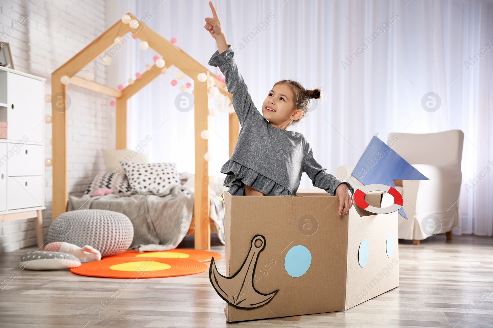 Photo of Cute little girl playing with cardboard boat in bedroom