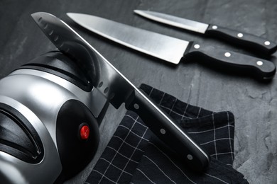Photo of Modern electrical sharpener and set of knives on black table, closeup