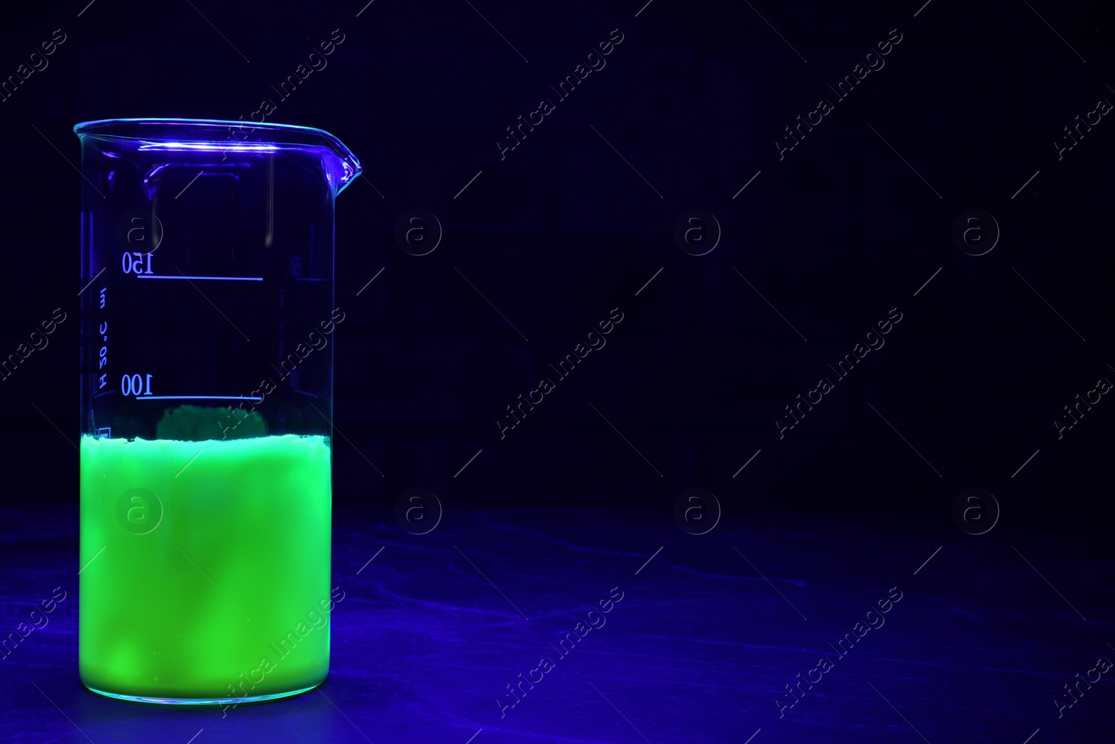 Photo of Laboratory beaker with luminous liquid on table against dark background, space for text