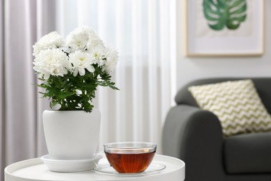 Photo of Beautiful chrysanthemum plant in flower pot and cup of tea on white table in room, space for text