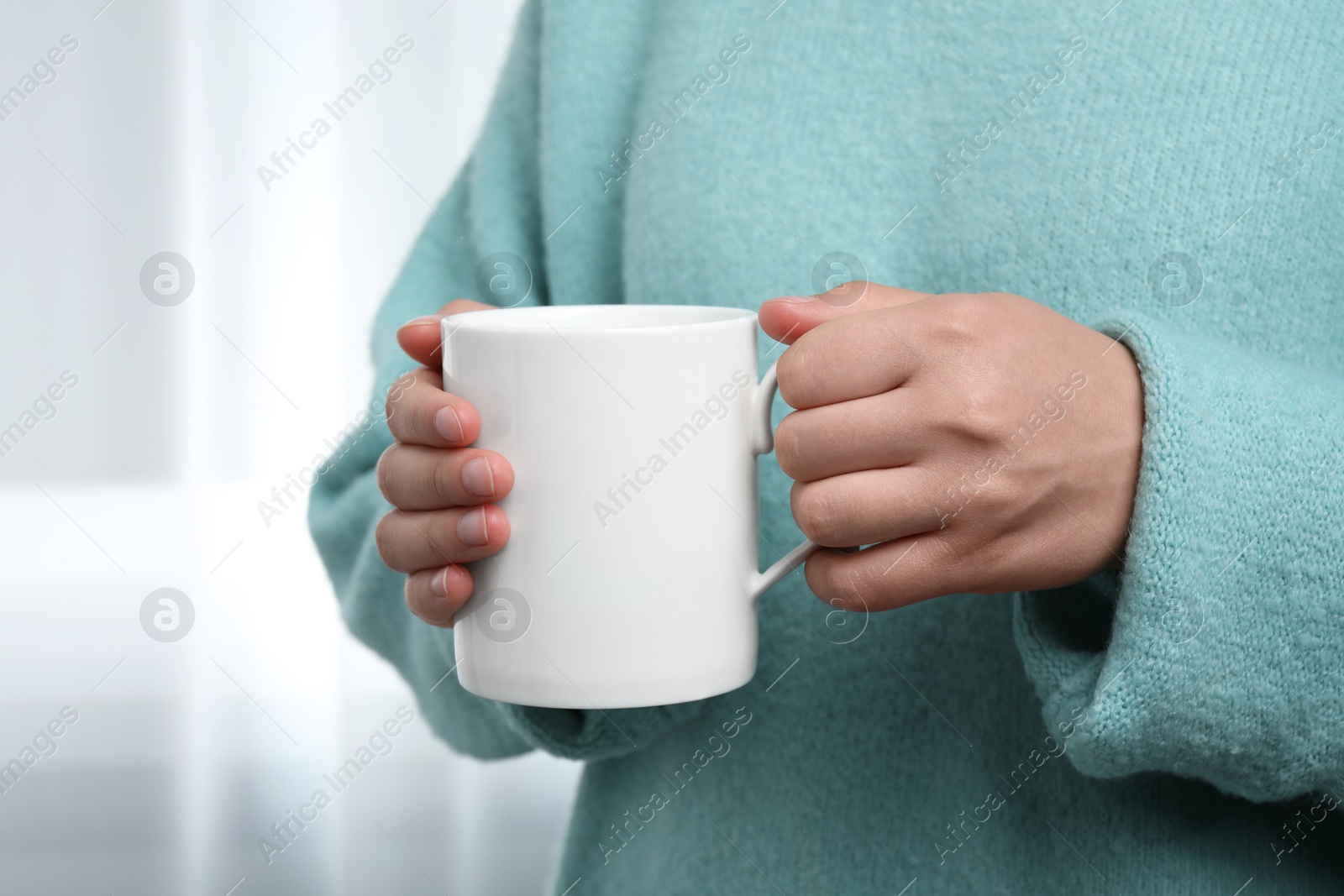 Photo of Woman holding white mug indoors, closeup. Mockup for design