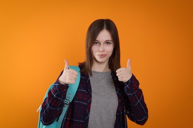 Photo of Portrait of cute teenage girl with backpack showing thumbs up on orange background