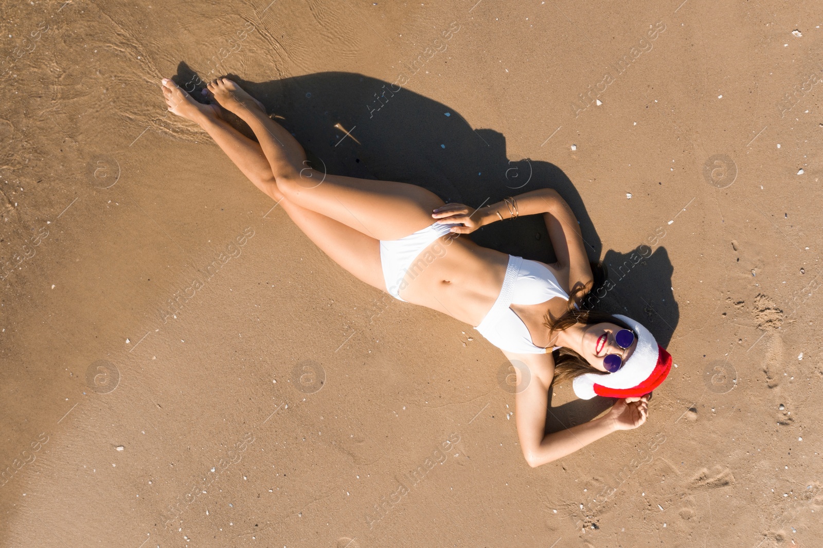 Image of Young woman wearing Santa hat and bikini on sunny beach, top view. Christmas vacation
