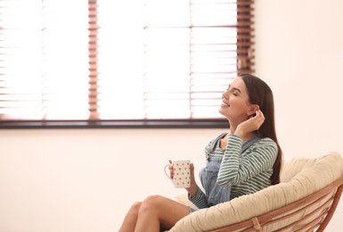 Photo of Young woman relaxing in papasan chair near window with blinds at home. Space for text