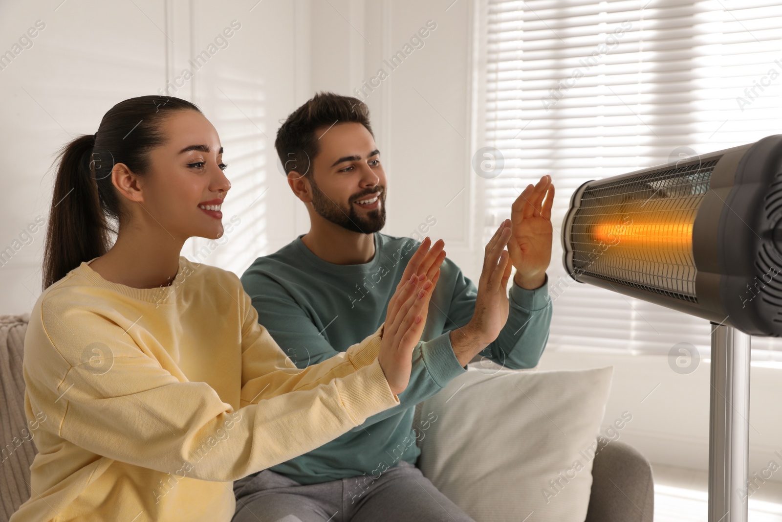 Photo of Young couple warming hands near electric heater at home