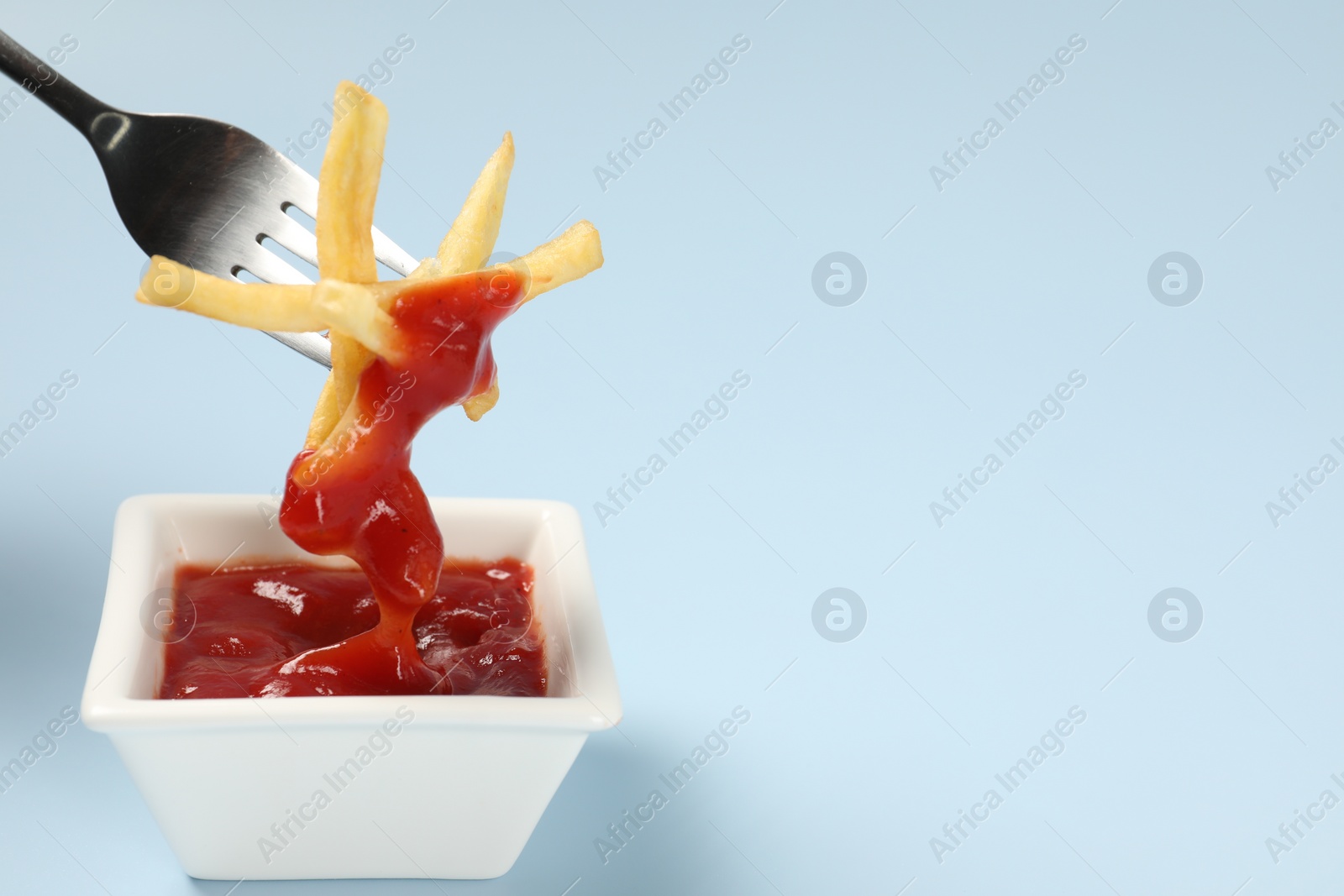 Photo of Dipping delicious french fries into ketchup on light blue background, closeup. Space for text