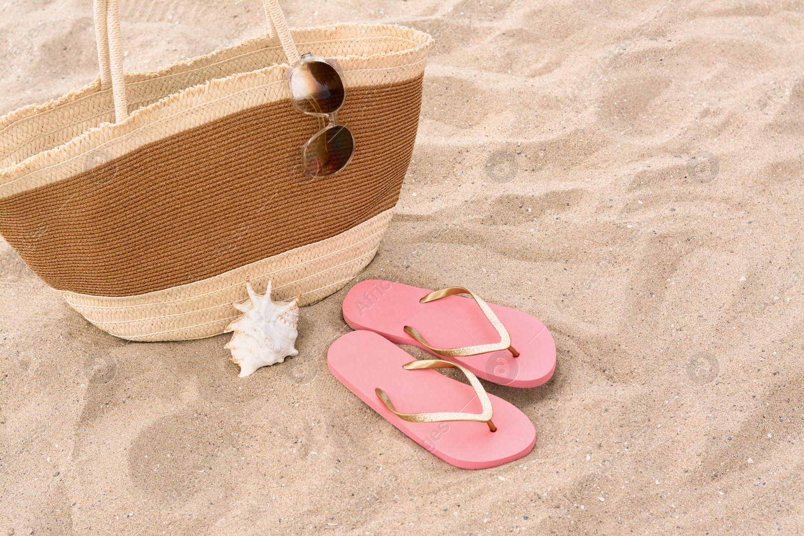 Photo of Stylish straw bag with sunglasses, flip flops and seashell on sand outdoors