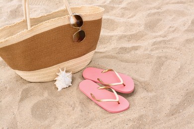 Stylish straw bag with sunglasses, flip flops and seashell on sand outdoors