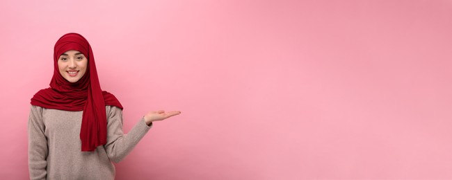 Image of Muslim woman in hijab pointing at something on pink background, space for text. Banner design