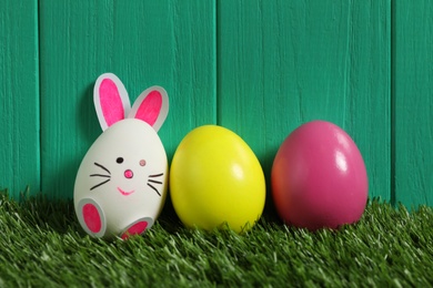 Photo of Bright eggs and white one as Easter bunny on green grass against wooden background