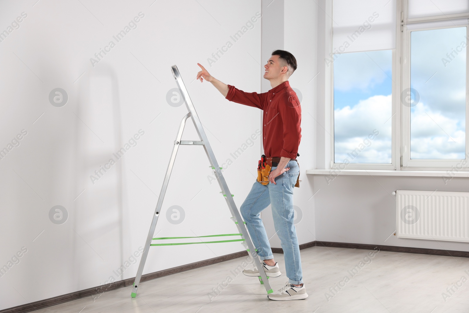 Photo of Young handyman with stepladder and tool belt working in room