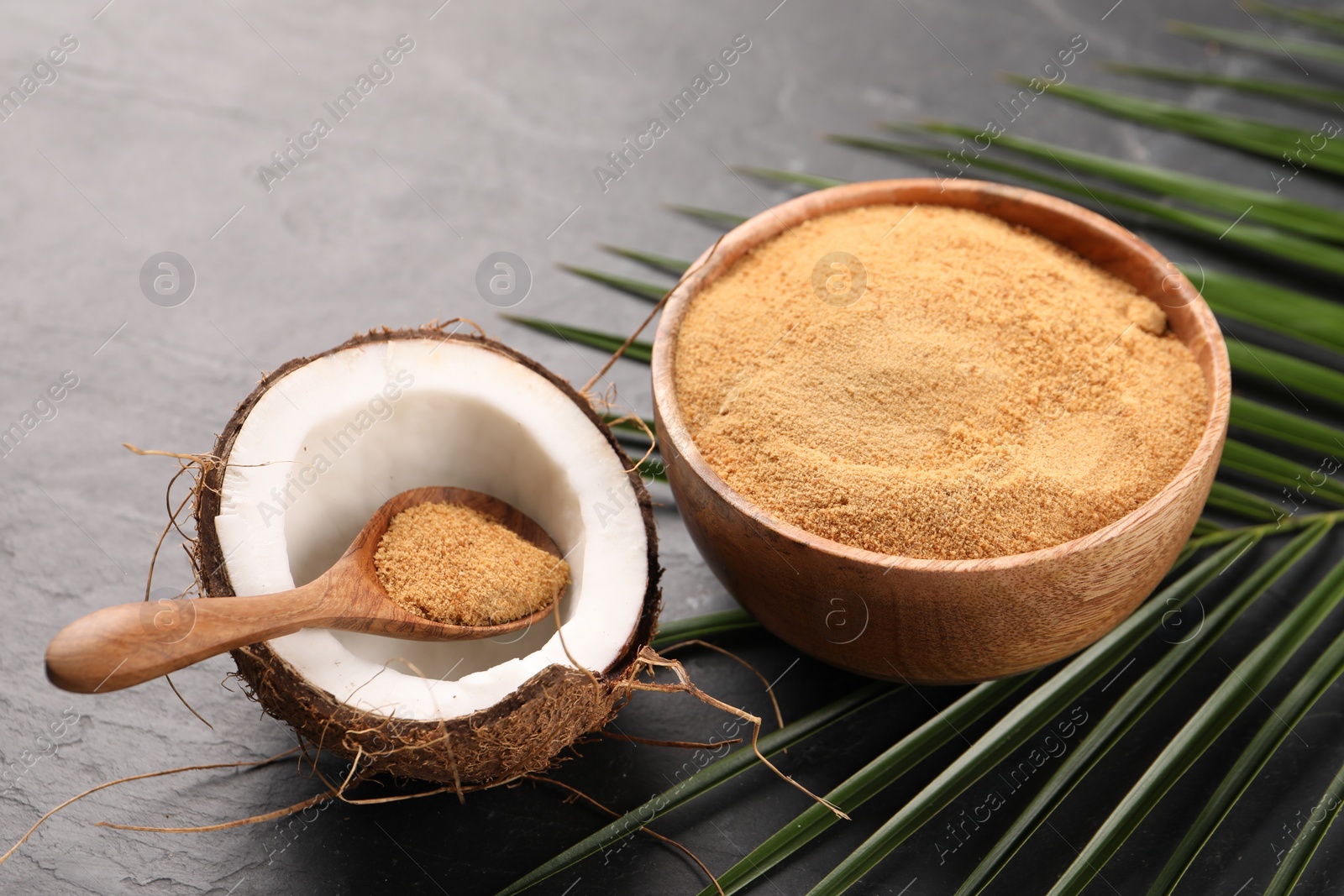 Photo of Spoon with coconut sugar, fruit, bowl and palm leaves on dark textured table, closeup