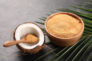 Photo of Spoon with coconut sugar, fruit, bowl and palm leaves on dark textured table, closeup