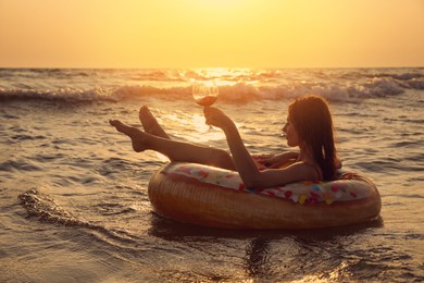 Woman with glass of wine and inflatable ring in sea at sunset