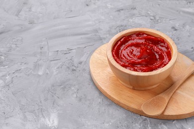 Photo of Organic ketchup in wooden bowl and spoon on grey textured table, space for text. Tomato sauce